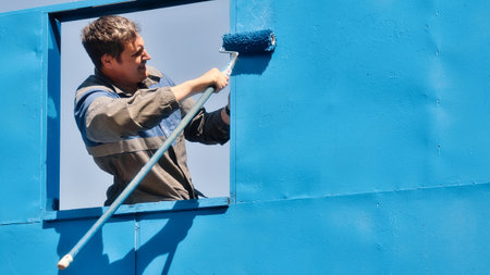 Professional painter in overalls paints metal wall of building with paint roller on summer day. Advertising background with copy space. Painting of metal surfaces. Authentic workflow.の写真素材