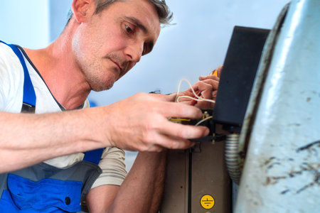Professional electrician at work. Electrician in work overalls repairs starter mechanism of unit or machine in factory indoors. Authentic workflow.の写真素材