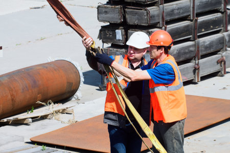 Two helmeted slingers attach load to slings outside on summer afternoon. Loading cargo. Real workers.の写真素材