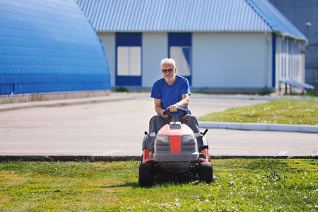 Elderly handyman gardener mows lawn on lawn mower tractor. Elderly man works as laborer. Mowing territory of industrial facility on summer day. Real workflow.の写真素材