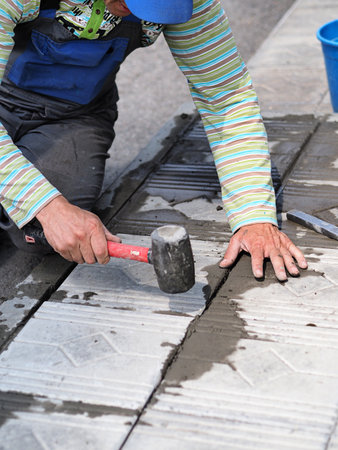 Tiler worker laying tiles on foundation of building outside. Hands of worker with hammer close-up. Construction worker laying paving slabs on summer day.の写真素材