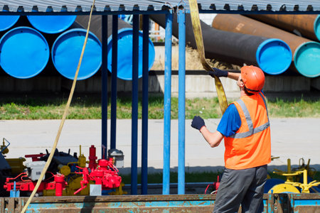 Slinger in hard hat and high-visibility vest unloads metal pipes for gasification on summer day in open warehouse outside. Real workflow.の写真素材