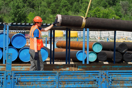 Slinger in hard hat and high-visibility vest unloads metal pipes for gasification on summer day in open warehouse outside. Real workflow.の写真素材