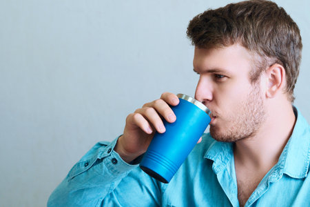 Young man drinks drink or medicine from thermo mug. Portrait of man from side on gray background. Drinks liquid from glass. copy space.の写真素材