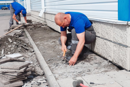 Elderly bricklayer lays tiles on foundation of building outside. Construction contractor lays out facing tiles of facade of production facility. Man lays tiles on summer day.の写真素材