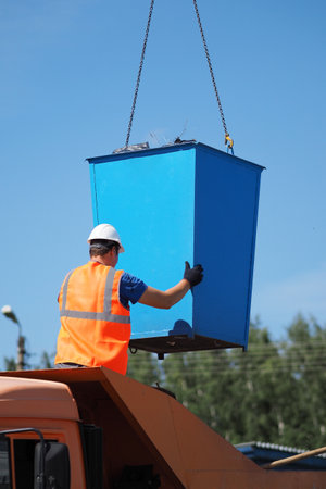 Handyman in protective vest and hard hat unloads garbage can into back of truck on summer day. Utilization of industrial waste. Working process.の写真素材