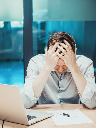 Office worker sits at table and holds his head in his hands. Depression and crisis in workplace....