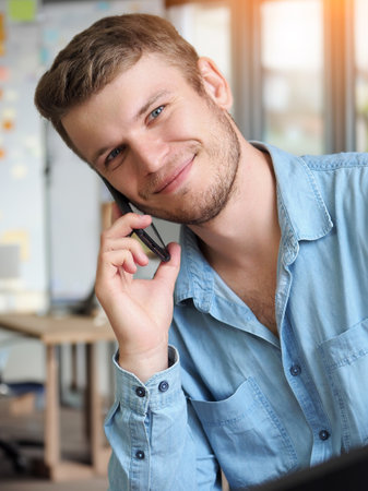 Handsome man is talking on phone and smiling. Operator takes phone call. Caucasian guy in denim shirt is sitting in office and talking on mobile phone. Positive emotions. Friendly support service.の写真素材