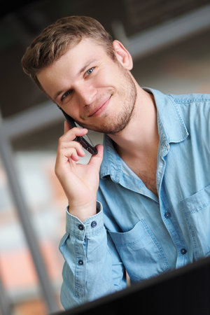 Handsome man is talking on phone and smiling. Operator takes phone call. Caucasian guy in denim shirt is sitting in office and talking on mobile phone. Positive emotions. Friendly support service.の写真素材