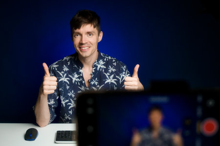 Happy blogger showing thumbs up to camera. Caucasian guy sits in home studio in front of phone camera and broadcasts live.の写真素材