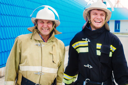 Two cheerful firefighters. Two firefighters in helmets and protective clothing stand outside on street and laugh cheerfully. Portrait of happy rescuers.の写真素材