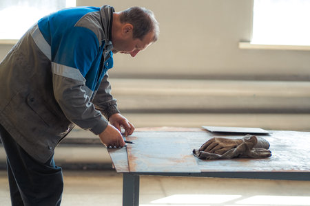 Worker in overalls works in workshop at workbench. Man marks metal sheet. Authentic portrait of worker at work.の写真素材