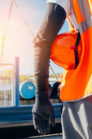 Builder or worker keeps hard hat on hand. Rear view from back. Slinger at construction site. Worker in overalls and construction vest.の写真素材