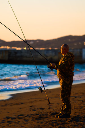 Elderly man fishes with fishing rod on ocean shore at sunset. Fisherman holds fishing rod in his hands and catches fish by sea. Leisure of pensioner. Hobby: fishing.の写真素材