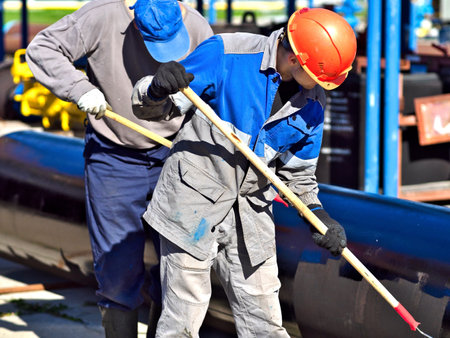 Two workers paint metal pipe black with paint rollers. Pipe treatment for corrosion protection. Metal paint. Two handymen paint gasification pipe on summer day.の写真素材