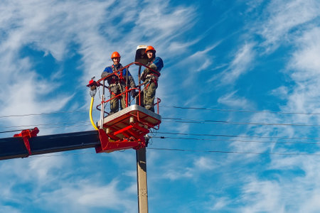 Two electricians from cradle of aerial platform or crane are repairing street lighting lamp. Professional electricians wearing helmets, overalls and insurance work at heights. View of workers from bottom up against sky. Authentic workflow.の写真素材
