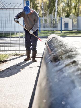 Man handyman paints large diameter metal pipe outside with paint roller on summer day. Protection of metal from corrosion.の写真素材