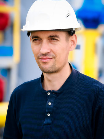 Portrait of Caucasian builder or gas industry engineer wearing white construction helmet. Confident looking working man aged 35-40 stands against background of yellow pipes. Reliable contractor.の写真素材