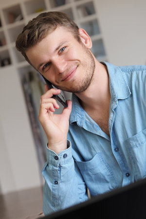 Handsome man is talking on phone and smiling. Operator takes phone call. Caucasian guy in denim shirt is sitting in office and talking on mobile phone. Positive emotions. Friendly support service.の写真素材