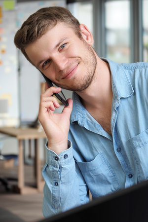 Handsome man is talking on phone and smiling. Operator takes phone call. Caucasian guy in denim shirt is sitting in office and talking on mobile phone. Positive emotions. Friendly support service.の写真素材