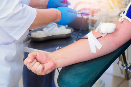 A donor donates blood at a blood transfusion station. Donors hand close-up. Healthcare and charity. A medical worker wearing a gown and gloves inserts a needle into a vein. World Donation Day.の写真素材