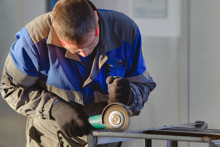 Man in overalls and safety glasses works with angle grinder in workshop. Worker grinds metal. working specialty. Blue collar. Daylight.の写真素材
