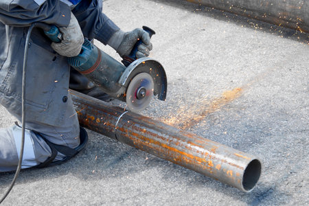 A worker wearing knee pads is cutting a large metal pipe with an angle grinder. The grinder is creating sparks and leaving a trail of metal shavings on the ground.の写真素材