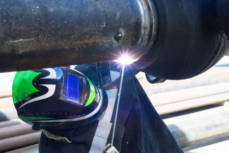 A welder, wearing a protective helmet and gloves, works on a large metal structure, sparks flying from the welding torch. The structure appears to be part of a larger project, with other pieces of metal visible in the background.の写真素材