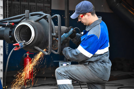 A worker in gray overalls and a blue cap works with industrial equipment, creating sparks. The workshop displays tools and safety equipment.の写真素材