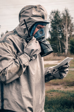 A person in full protective clothing and a gas mask is seen using a radio outdoors, with a document in hand, indicating a field operation or emergency situation.の写真素材