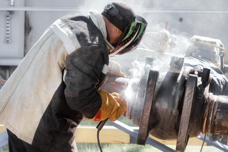 Worker welding a large industrial pipe joint in an outdoor setting, wearing necessary protective clothing and equipment to ensure safety during the high-temperature process.の写真素材