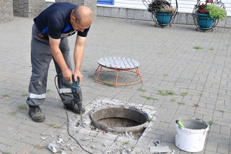A worker uses a handheld drill to repair a manhole cover in a city area. The scene features a circular opening in the pavement surrounded by debris, with tools and materials nearby. Green plants add a touch of nature.の写真素材