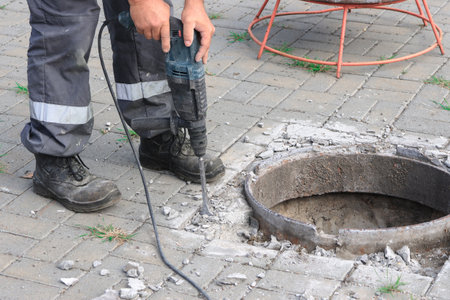 Worker in protective gear uses a power drill to break concrete around a circular opening in pavement, ensuring safety and precision in a construction site environment.の写真素材
