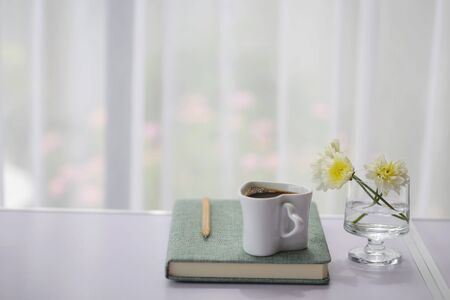 Coffee cup and flowers in glass with diary notebooks and pencil on table in front of see through...