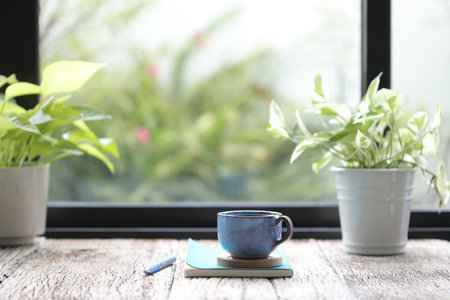 Blue clay coffee cup with green plant pot and notebook with blue pencil on wooden table infront of windowsの写真素材