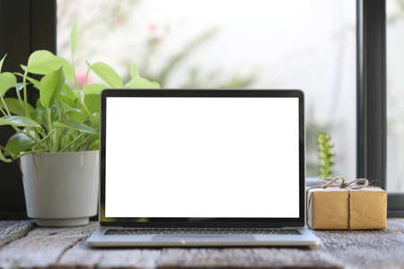 Laptop mock up white blank screen with  gift box and plant pot on wooden table in front of windows