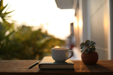White cup and notebook on wooden table under sunset lightの写真素材