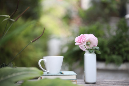 white coffee cup with pink rose in ceramic vase and notebooks on weathered wooden table at outdoorの写真素材