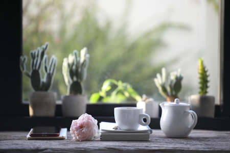 White coffee cup and ceramic teapot and pink rose with notebook on wooden table and cactus pot in front of windowの写真素材