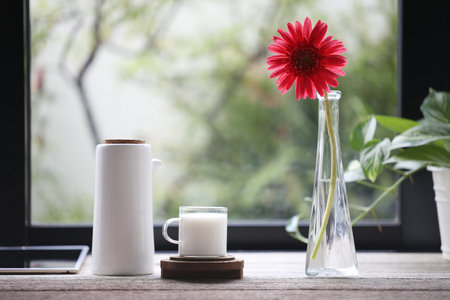 a glass cup of milk and pitcher pot and flower in glass vase on wooden table in front of windowの写真素材