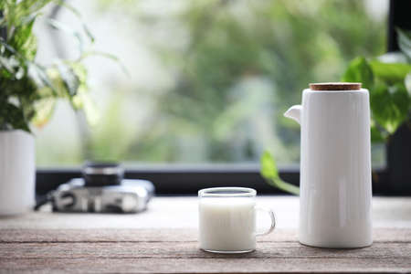 a glass cup of milk and pitcher pot and on wooden table in front of windowの写真素材