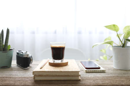 Coffee with crema in a a glass cup and notebooks and phone on wooden table with plants