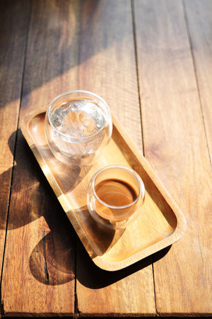 Hot coffee and glass of ice on wooden tray under sunlightの写真素材
