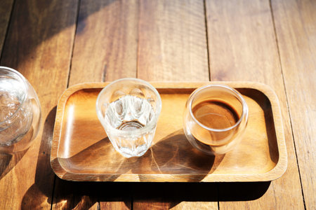 Hot coffee and glass of ice on wooden tray under sunlightの写真素材