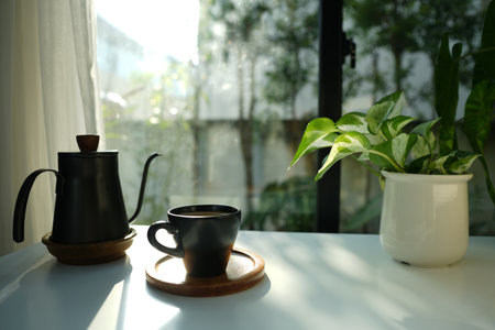 Black coffee cup and pot with potted plant on white table, by the windowの写真素材