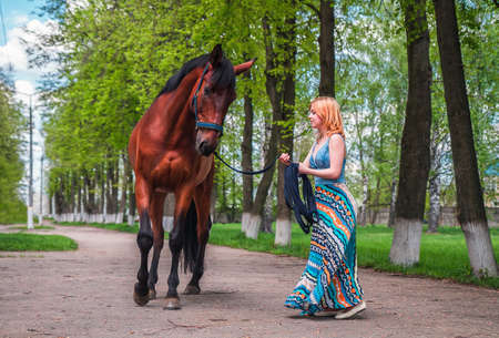 A young blond woman in a long blue dress leads the brown horse on the track along the high treeの写真素材