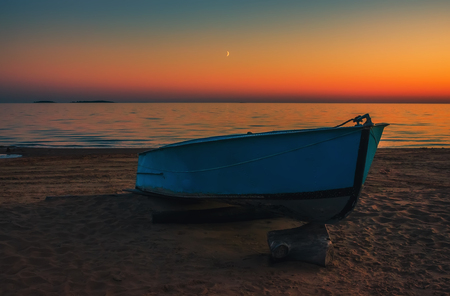 Blue metal boat on the sandy beach against the backdrop of an orange night sky reflected in the sea.の写真素材