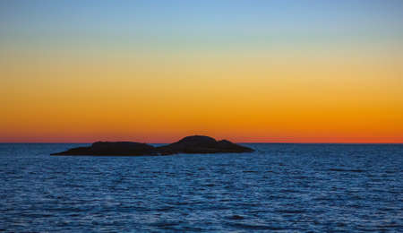 Silhouette of a small island on a background of orange sky horizon separated from the blue surface of the water.の写真素材