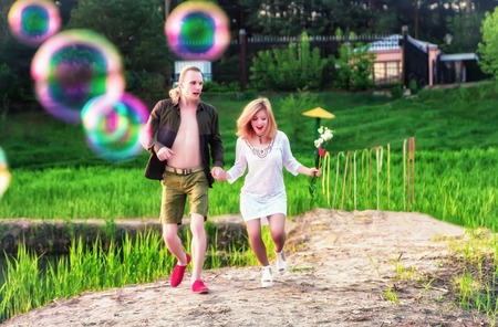 Young happy couple running outdoors among green grass and soap bubbles at sunny summer day....