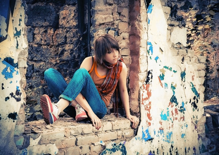 Young girl in an orange shirt and blue jeans sitting with a thoughtful look at the ruins of an abandoned brick building with graffiti on the walls. Selective focus.の写真素材